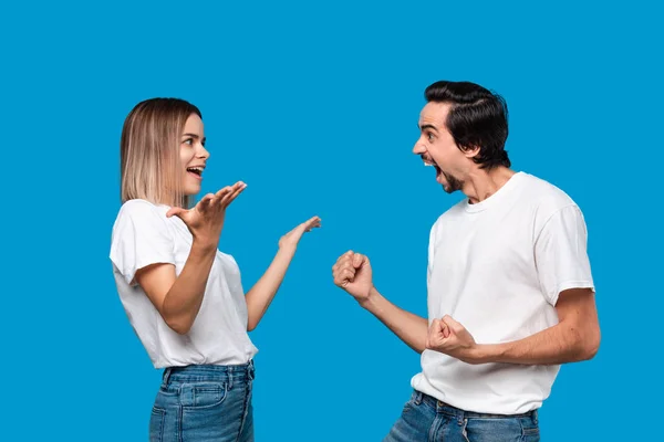 Emotional couple expressing gesture of success. Young blond woman and bearded man with mustaches in white tees and blue jeans are winner. Models standing isolated over blue background.
