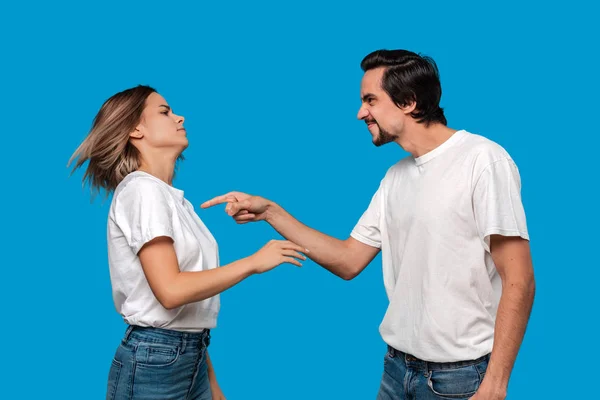 Pareja de una joven rubia y un hombre barbudo morena con bigotes en camisetas blancas y vaqueros azules peleando de pie aislado sobre fondo azul . — Foto de Stock