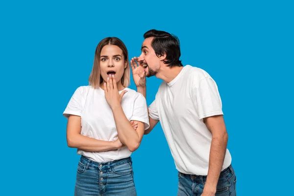 Hombre barbudo con bigotes en camiseta blanca le cuenta el secreto a una joven rubia con camisetas blancas y pantalones vaqueros azules de pie aislados sobre fondo azul. Concepto de narración secreta . — Foto de Stock