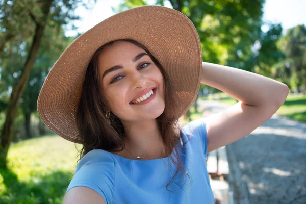 Улыбающаяся молодая женщина в синем платье и шляпе с широкими закрылками сидит в парке и делает селфи со смартфоном . — стоковое фото