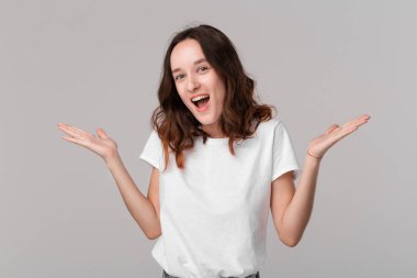 Beyaz bir tee heyecanlı esmer kadın gri arka plan üzerinde izole ayakta sürpriz duygu ifade omuzlarını silkme. Sürpriz duygu.
