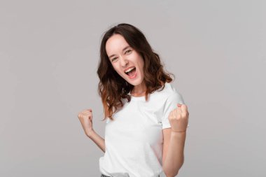 Gri arka plan üzerinde izole ayakta başarı duygularını ifade beyaz bir tee heyecanlı esmer kadın. Başarı duygusu.