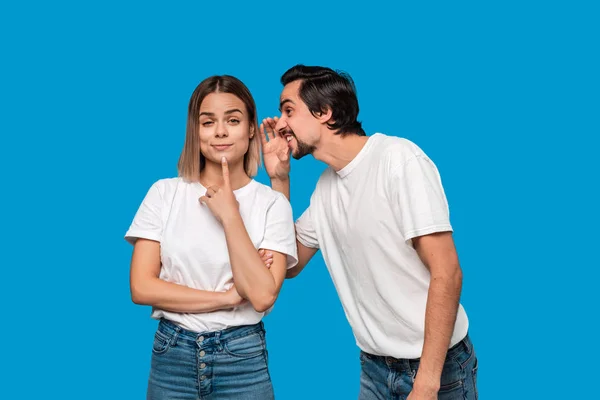 Bearded man with mustaches in white t-shirt tells secret to a young blond girl in white t-shirts and blue jeans standing isolated over blue background. Concept of secret telling.