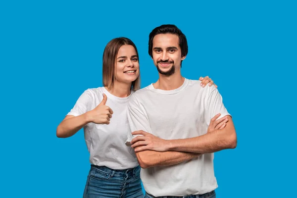 Happy couple of a young blond woman and brunet bearded man with mustaches in white t-shirts and blue jeans posing isolated over blue background. Concept of an ideal couple. Models holding thumbs up