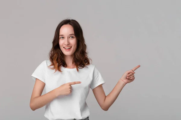 Hübsche brünette Frau in einem weißen T-Shirt, die auf den leeren Raum in einer rechten Seite eines Rahmens zeigt, der isoliert vor grauem Hintergrund steht. — Stockfoto