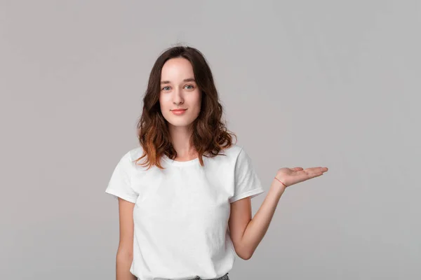 Mulher morena bonita em um tee branco segurando palma de uma mão com espaço vazio para a colocação do produto de pé isolado sobre fundo cinza . — Fotografia de Stock