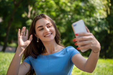 Mavi elbiseli mutlu genç kadın parkta oturuyor ve akıllı telefonla selfie çekiyor.