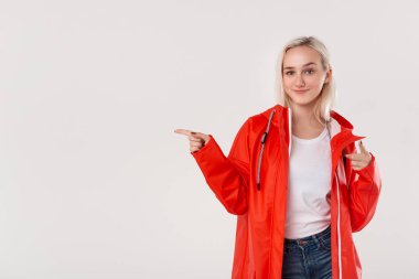 Beyaz arka plan üzerinde izole boş alanda parmak ile işaret kapüşonlu kırmızı yağmurluk içinde Gülümseyen sarışın kız. Yağmur yağmaya başladı. Reklam yeri