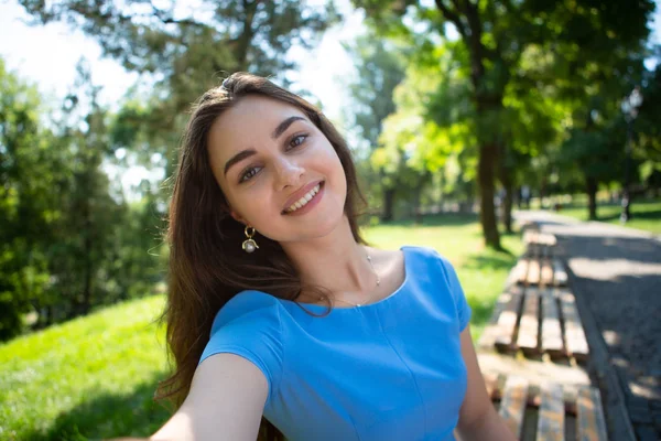 Mavi elbiseli mutlu genç kadın parkta oturuyor ve akıllı telefonla selfie çekiyor.