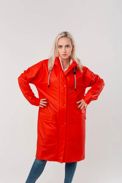 Mooi blond meisje in een rode regenjas staande met armen a-Kimbo geïsoleerd over witte achtergrond. Maak je klaar voor slecht weer. — Stockfoto