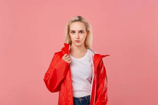 Linda chica rubia con un impermeable rojo y una camiseta blanca de pie aislada sobre fondo rosa. Prepárate para el mal tiempo . — Foto de Stock