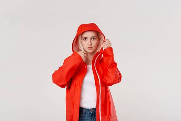Blond girl gets on hood of a red raincoat isolated over white background. It is starting to rain.