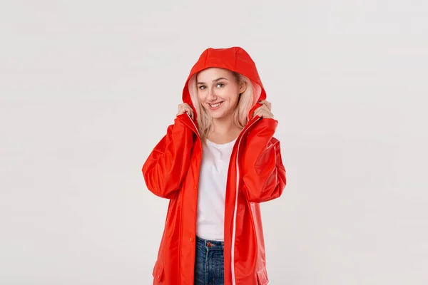Chica rubia sonriente con un impermeable rojo con capucha escondida de la lluvia aislada sobre fondo blanco. Está empezando a llover. . —  Fotos de Stock