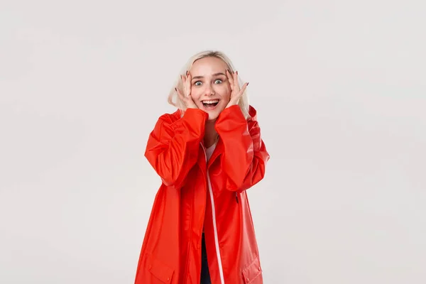 Chica rubia excitada en un impermeable rojo con capucha escondida detrás de un plano de manos expresando emoción de felicidad aislada sobre fondo blanco. Prepárate para el tiempo lluvioso . —  Fotos de Stock