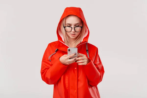 Mulher loira bonita em uma capa de chuva vermelha com um smartphone de verificação de capuz para notificações isoladas sobre fundo branco. Indo para um passeio em um dia frio chuvoso — Fotografia de Stock