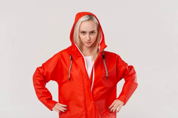 白い背景の上に孤立した腕で立っている赤いレインコートのかわいいブロンドの女の子。悪天候の準備はできていますか? — ストック写真