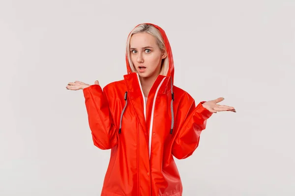 Blond girl in a red raincoat with hood expressing emotion of indignation  because of rainy weather isolated over white background. Get ready for bad weather.