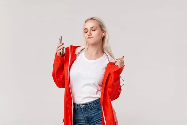 Linda chica rubia con impermeable rojo y camiseta blanca bailando escuchando música con auriculares inalámbricos. Concepto de disfrutar de la vida . — Foto de Stock