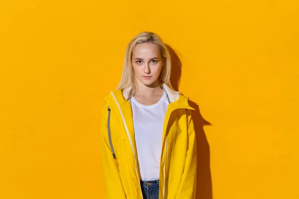 Linda chica rubia con un impermeable amarillo y una camiseta blanca de pie aislada sobre fondo amarillo. Prepárate para el mal tiempo . — Foto de Stock