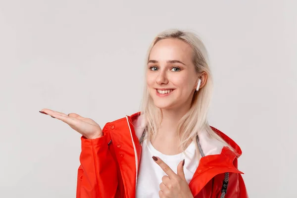 白い背景の上に隔離された手のひらの上の空のスペースに指を指すフード付きの赤いレインコートを着たワイヤレスイヤホンを持つ笑顔のブロンドの女の子。広告の場所 — ストック写真