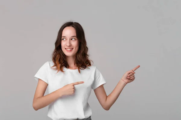 Hübsche brünette Frau in einem weißen T-Shirt, die auf den leeren Raum in einer rechten Seite eines Rahmens zeigt, der isoliert vor grauem Hintergrund steht. Kopierraum der Werbung. — Stockfoto