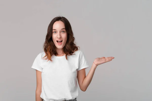 Hübsche brünette Frau in einem weißen T-Shirt, das eine Handfläche mit leerem Raum für Produktplatzierung hält und isoliert vor grauem Hintergrund steht. Kopierraum der Werbung. — Stockfoto