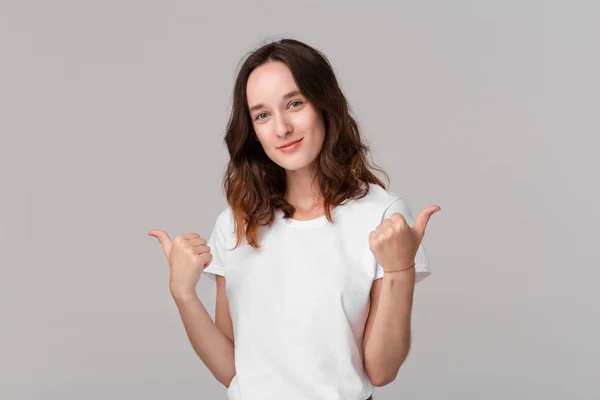 Mooie brunette vrouw in wit t-shirt met duimen omhoog uitdrukken positieve evaluatie permanent geïsoleerd over grijze achtergrond. Ik vind het leuk! — Stockfoto
