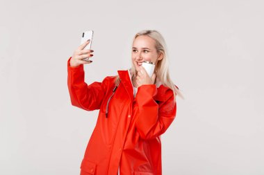Kırmızı yağmurluk lu güzel sarışın kadın, beyaz arka plan üzerinde izole sıcak içecek ile beyaz termal kupa tutan akıllı telefon ile selfie yapıyor. Yağmurlu bir soğuk günde yürüyüşe çıkmak