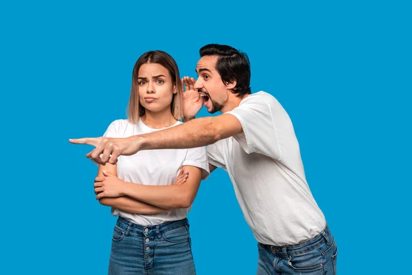 Bearded man with mustaches in white t-shirt tells secret to a young blond girl in white t-shirts and blue jeans standing isolated over blue background. Woman looks surprised. Secret telling.