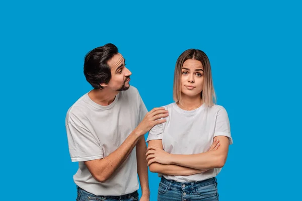 Brunet bearded man with mustaches in white t-shirt and blue jeans asking his girlfriend for forgiveness isolated over blue background. Concept of guilt. — Stock Photo, Image