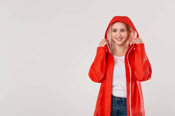 笑顔のブロンドの女の子は、白い背景の上に隔離された赤いレインコートのフードに乗ります。雨が降り出し始めている. — ストック写真