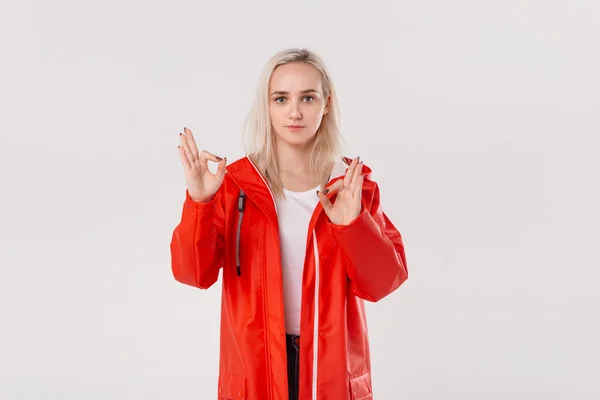 Linda chica rubia con un impermeable rojo con capucha que muestra signo OK aislado sobre fondo blanco. Prepárate para el tiempo lluvioso . —  Fotos de Stock