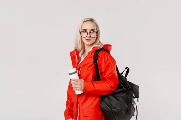 Kırmızı yağmurluk ve gözlük lü sarışın kız soğuk mevsimde siyah sırt çantası ve beyaz termal kupa ile seyahat. Yağmur mevsimi. — Stok fotoğraf