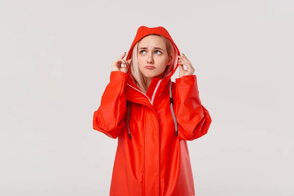 Chica rubia con un impermeable rojo con una capucha comprobando si la lluvia ha comenzado a estar aislada sobre fondo blanco. ¿Estás listo para el mal tiempo frío ? —  Fotos de Stock