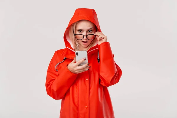 Mujer bastante rubia en un impermeable rojo con una capucha parece sorprendido comprobar teléfono inteligente para las notificaciones aisladas sobre fondo blanco. Voy a dar un paseo en un día frío lluvioso —  Fotos de Stock