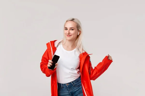 Linda chica rubia con impermeable rojo y camiseta blanca bailando escuchando música con auriculares inalámbricos. Concepto de disfrutar de la vida . — Foto de Stock