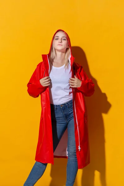 Nätt blond flicka i en röd regnrock och vit t-shirt stående isolerad över gul bakgrund. Vara ljus i dåligt väder. — Stockfoto