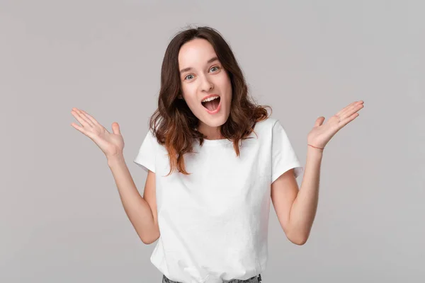 Opgewonden brunette vrouw in een witte tee afschudden haar schouders uitdrukken emotie van verrassing staande geïsoleerd over grijze achtergrond. Emotie van verrassing. — Stockfoto