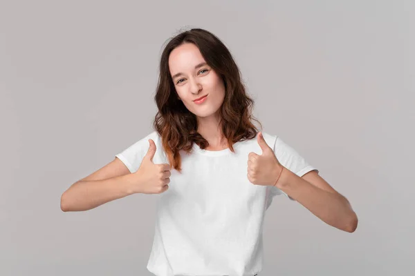 Mooie brunette vrouw in wit t-shirt met duimen omhoog uitdrukken positieve evaluatie permanent geïsoleerd over grijze achtergrond. Ik vind het leuk! — Stockfoto