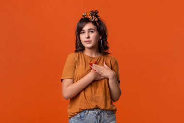 Bir t-shirt ve güzel kafa bandı güzel esmer kadın turuncu arka plan üzerinde izole ayakta samimiyet ifade kalp ellerini koyarak. Sencere duygular kavramı