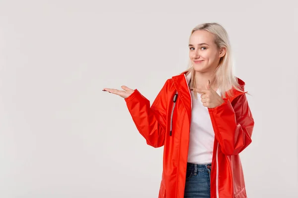 Linda chica rubia con impermeable rojo con capucha apuntando con el dedo al espacio vacío en una palma de una mano aislada sobre fondo blanco. Lugar para la publicidad — Foto de Stock