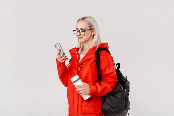 Menina loira em uma capa de chuva vermelha e óculos viajando com mochila preta e caneca térmica branca na estação fria. Temporada chuvosa . — Fotografia de Stock