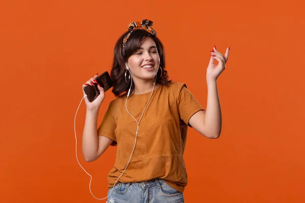 Bir t-shirt ve güzel kafa bandı turuncu arka plan üzerinde izole kablolu kulaklıklar ile müzik dinlerken dans Güzel esmer kadın. Hayatın tadını çıkarmak