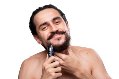 Bıyıklı yakışıklı adam beyaz arka plan üzerinde izole çıplak ayakta sakalını kesmek için elektrikli tıraş makinesi kullanır. Sabah tedavi ve tıraş kavramı. Sakalını kesme zamanı. Sabah rutini