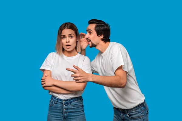 Beyaz t-shirt bıyıklı sakallı adam beyaz t-shirt ve mavi kot mavi arka plan üzerinde izole ayakta genç bir sarışın kız için gizli söyler. Kadın şaşırmış görünüyor. Gizli anlatım.
