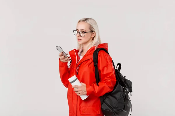 Fille blonde dans un imperméable rouge et des lunettes voyageant avec sac à dos noir et tasse thermique blanche en saison froide. Saison des pluies . — Photo