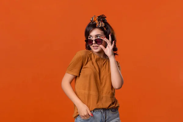 Kaunis brunette nainen t-paita ja kaunis otsanauha ilmaista tunteita yllätys tilalla aurinkolasit seisoo eristetty oranssi tausta . — kuvapankkivalokuva