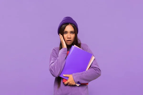 Adolescente afroamericano bastante en un sombrero y suéter sosteniendo carpetas y expresando emoción de sorpresa de pie aislado sobre fondo púrpura. Concepto de educación — Foto de Stock