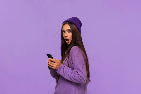 Pretty african-american girl in a hat and sweater holding smartphone and expressing emotion of surprise standing isolated over purple background. — Stock Photo, Image