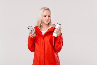 Kırmızı yağmurluklu güzel sarışın kadın elinde beyaz termal bardakla, beyaz arka planda izole bir şekilde duran sıcak bir içecek ve akıllı telefon tutuyor. Yağmurlu ve soğuk bir günde yürüyüşe çıkıyorum.